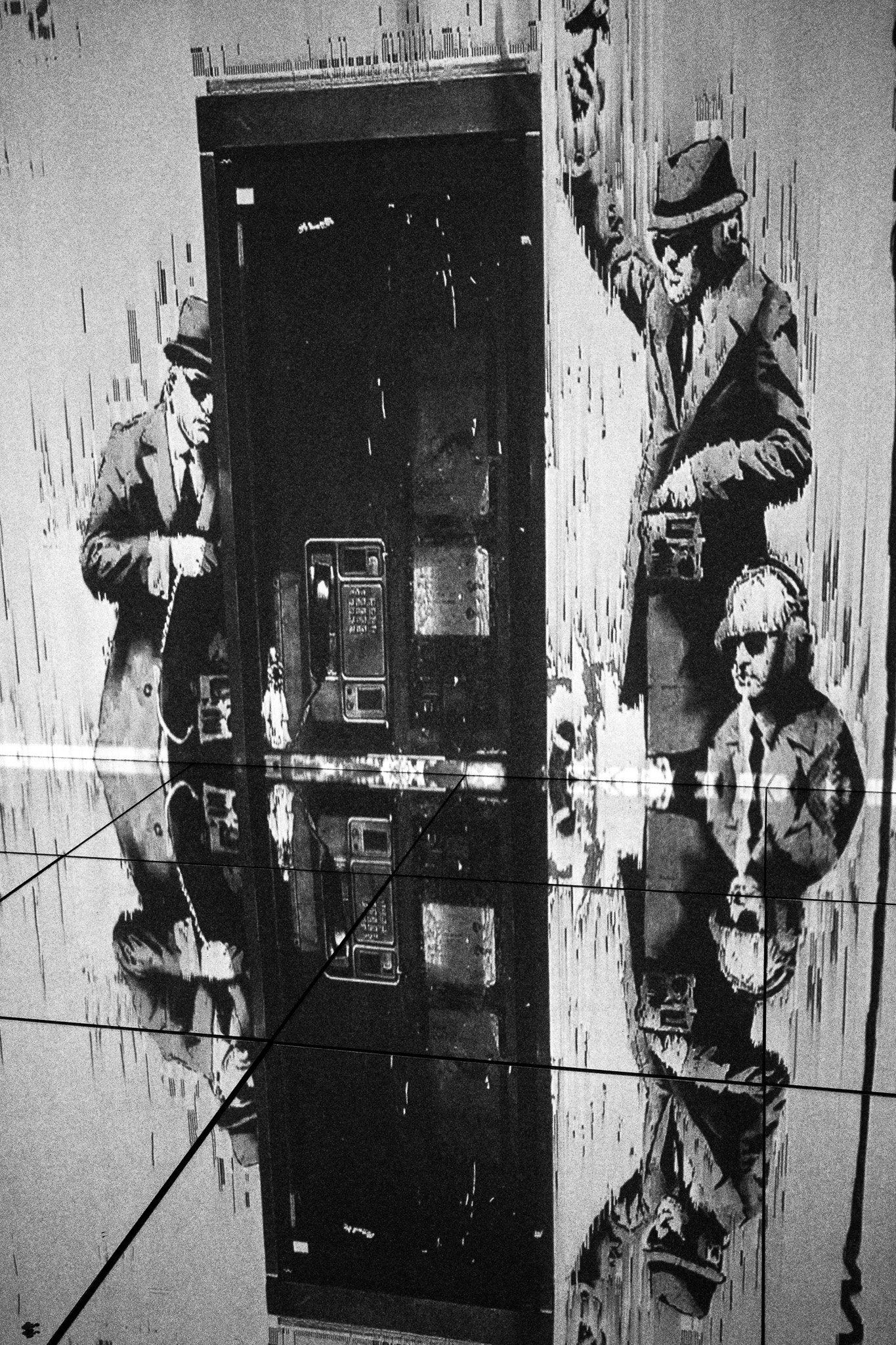 The image features a striking black-and-white mural depicting two men in suits, one seemingly engaged with a payphone, while the other stands confidently, possibly holding a camera. A young boy in a similar style is included, wearing dark sunglasses and looking toward the viewer. The scene is set against a textured wall that appears to be peeling, adding depth and a sense of urban decay. The reflection on a shiny, tiled floor amplifies the visual impact, drawing attention to the characters and their actions.
