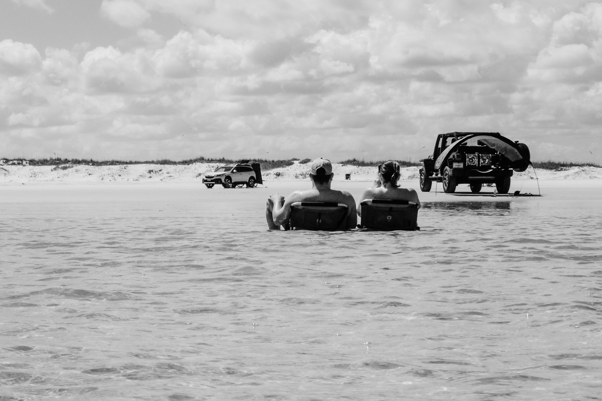 The image captures a serene beach scene in black and white, where a couple sits in beach chairs partially submerged in shallow water, enjoying a moment of relaxation. In the background, two vehicles are parked on the sandy shore, with one featuring a kayak or canoe secured on its roof. The sky is dotted with fluffy clouds, adding a tranquil ambiance to the setting, while the gentle ripples of the water emphasize the relaxed atmosphere.