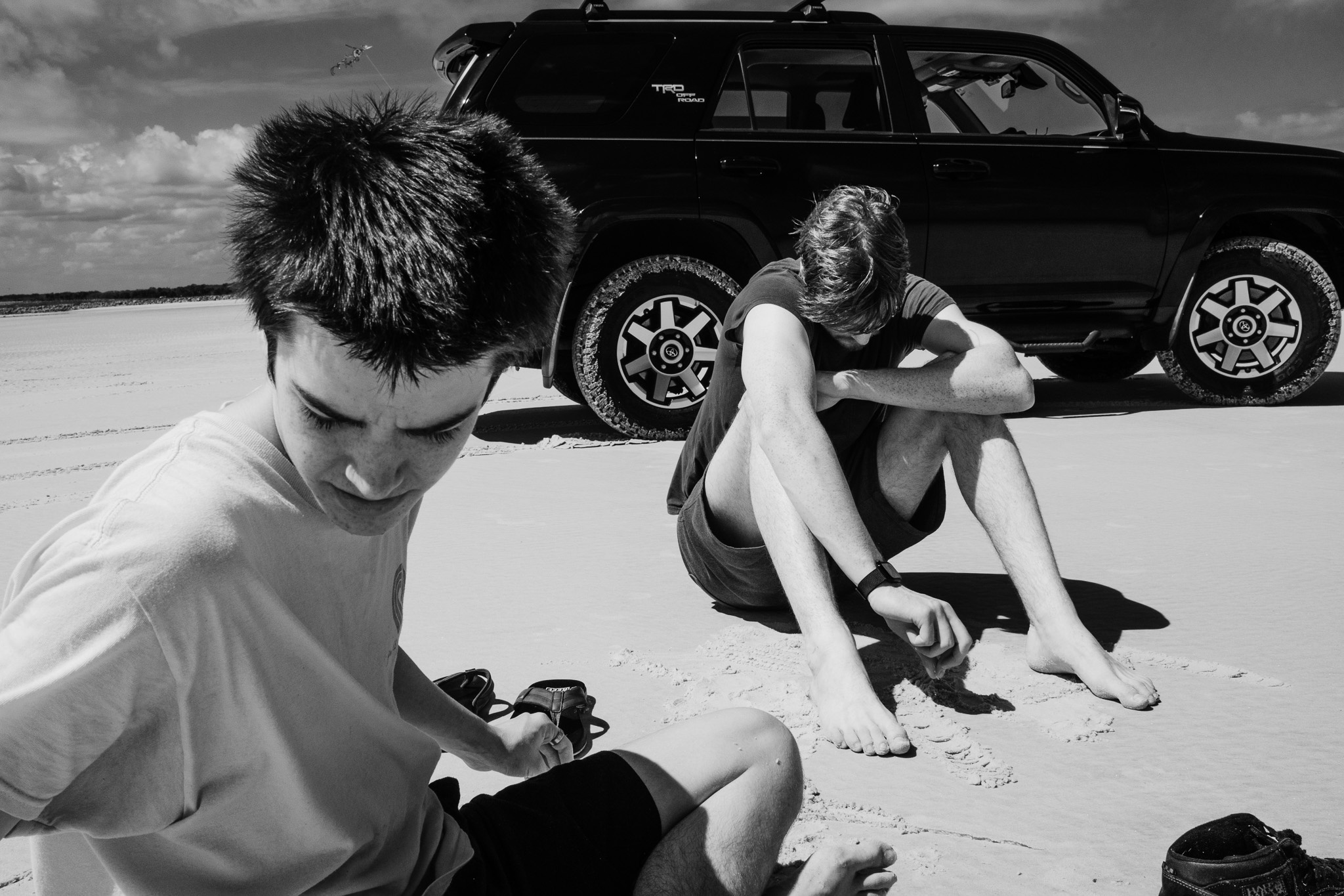 The image captures two young men sitting on a sandy beach under a bright sky, with a black SUV parked nearby. One boy, wearing a light-colored shirt, appears to be focused on the sand, while the other, in a dark shirt, leans forward with his arms crossed, seemingly deep in thought. The contrast of the black and white tones emphasizes the textures of the sand and their expressions, creating a contemplative atmosphere in this outdoor setting.