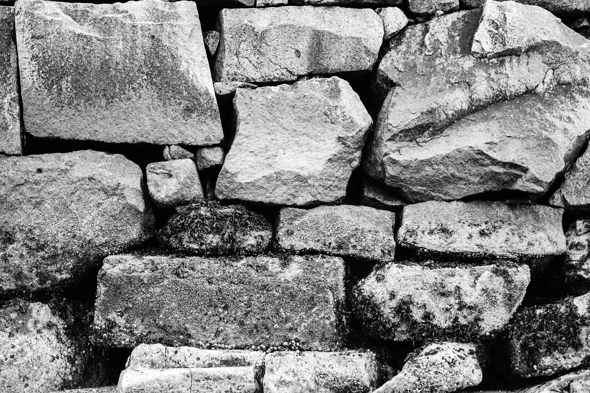 The image features a textured stone wall composed of various sizes of rough, gray stones, some of which are stacked neatly while others appear more haphazardly arranged. The stones display a weathered appearance, with moss and lichen growth in some areas, suggesting age and exposure to the elements. The monochromatic color scheme enhances the ruggedness of the materials and emphasizes the intricate patterns formed by the stones, creating a sense of natural beauty in the structure.