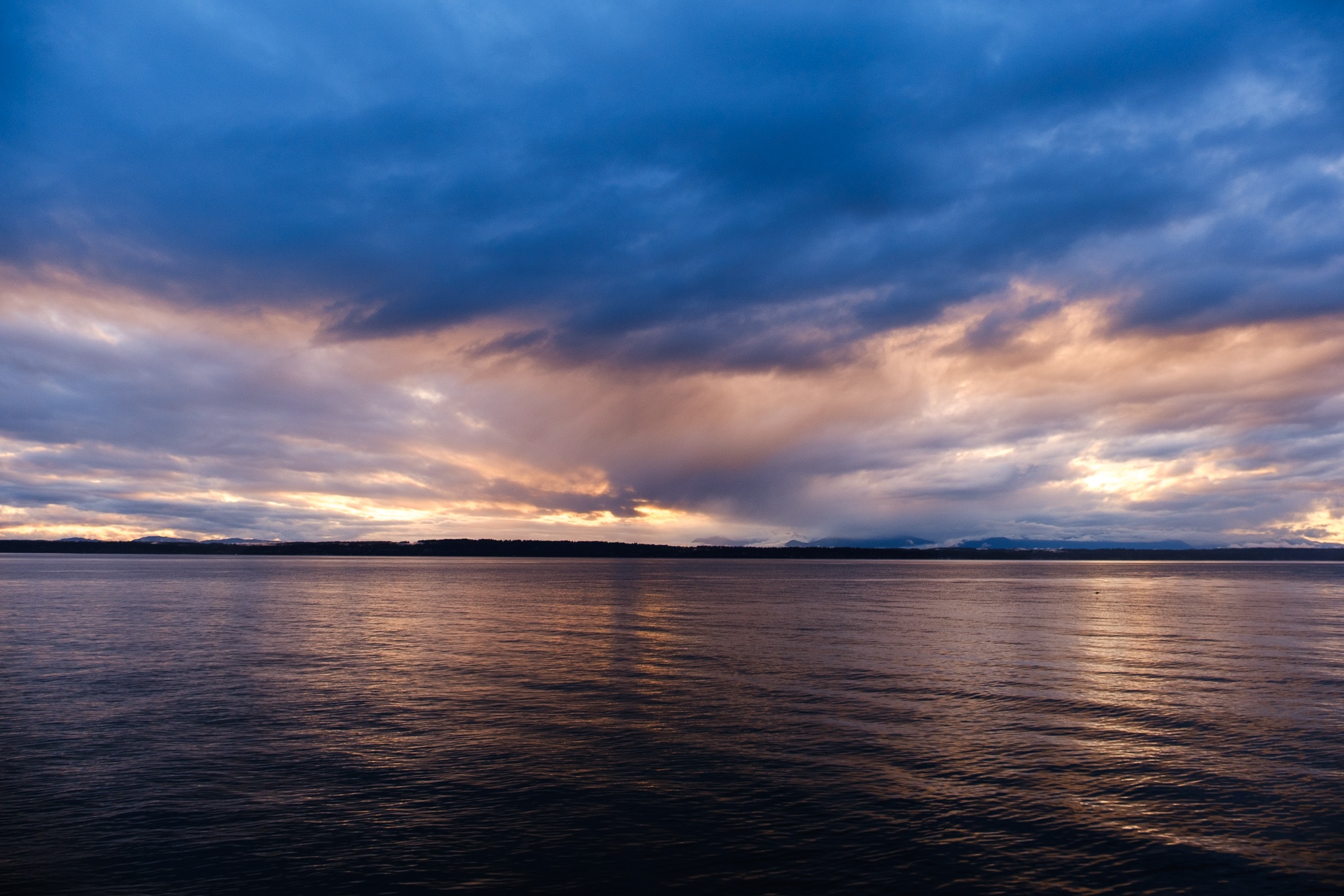 The image captures a serene waterfront scene during twilight, showcasing a tranquil body of water reflecting the soft hues of a cloudy sky. The clouds range from deep blues to warm oranges and pinks, suggesting a sunset atmosphere. In the background, a dark silhouette of distant hills adds depth to the composition, while the smooth surface of the water enhances the overall calmness of the setting. The interplay of light and shadow creates a visually striking contrast, inviting a sense of peace and contemplation.