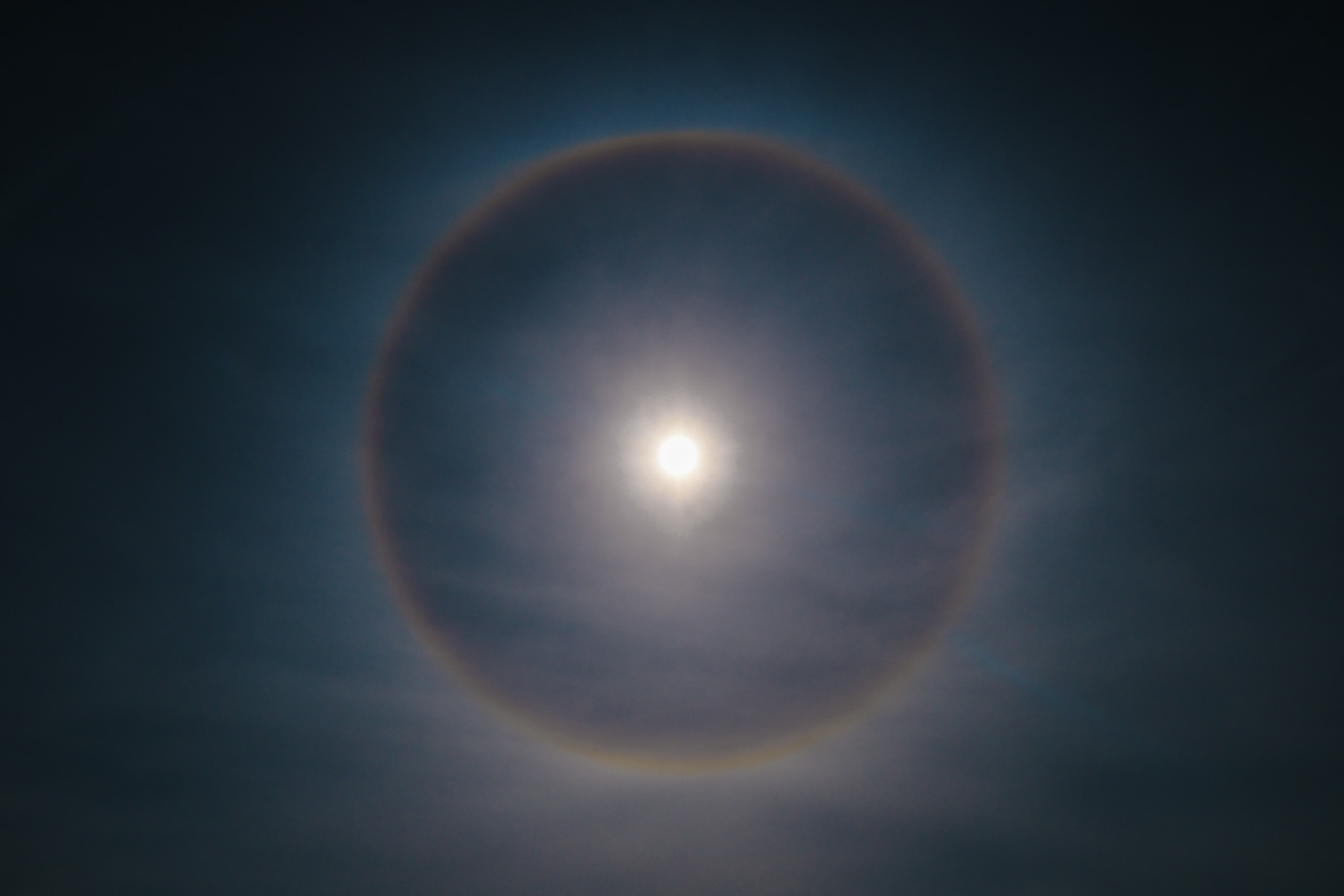 The image captures a striking atmospheric phenomenon featuring a bright sun at the center, surrounded by a vivid halo or ring of light. The halo displays a gradient of colors, fading from bright whites and yellows near the sun to deeper blues and purples at the edges. The background is a soft blend of dark sky hues, creating a dramatic contrast that enhances the brilliance of the halo. This celestial display suggests a serene yet awe-inspiring moment in nature, emphasizing the beauty and wonder of the sky.