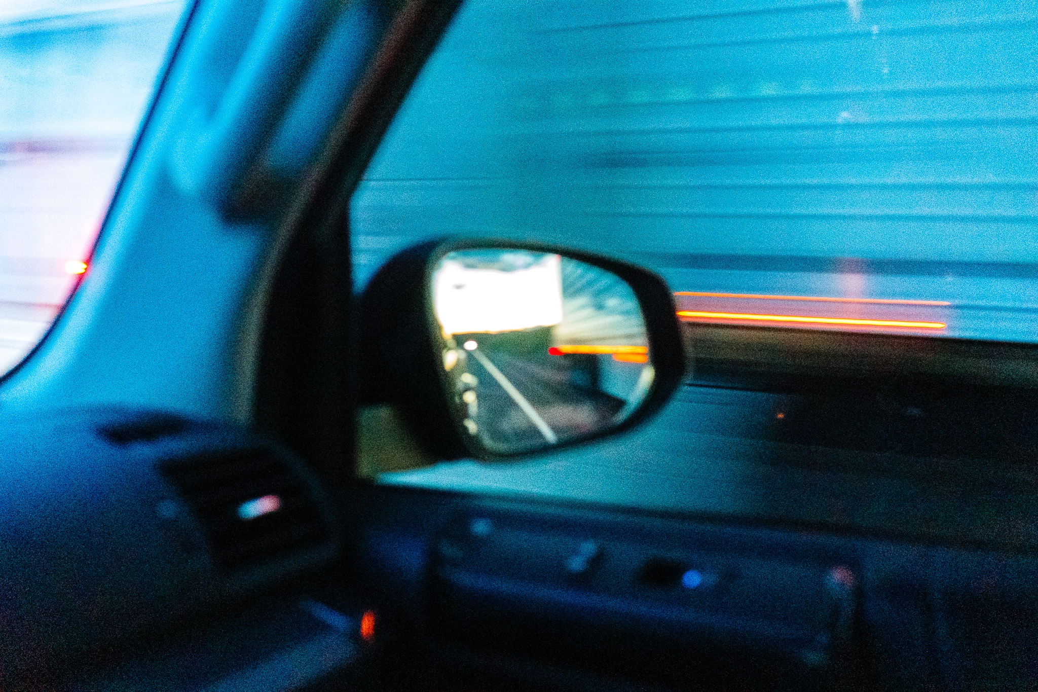 The image captures the view from inside a vehicle, focusing on the side mirror that reflects a blurred landscape of moving lights and colors, suggesting high speed. The interior of the car is partially visible, with a dark dashboard and air vent. The background features streaks of light, likely from other vehicles or streetlights, creating a dynamic sense of motion. The overall color palette is cool, dominated by blue tones, enhancing the feeling of speed and movement as the vehicle travels along a road.
