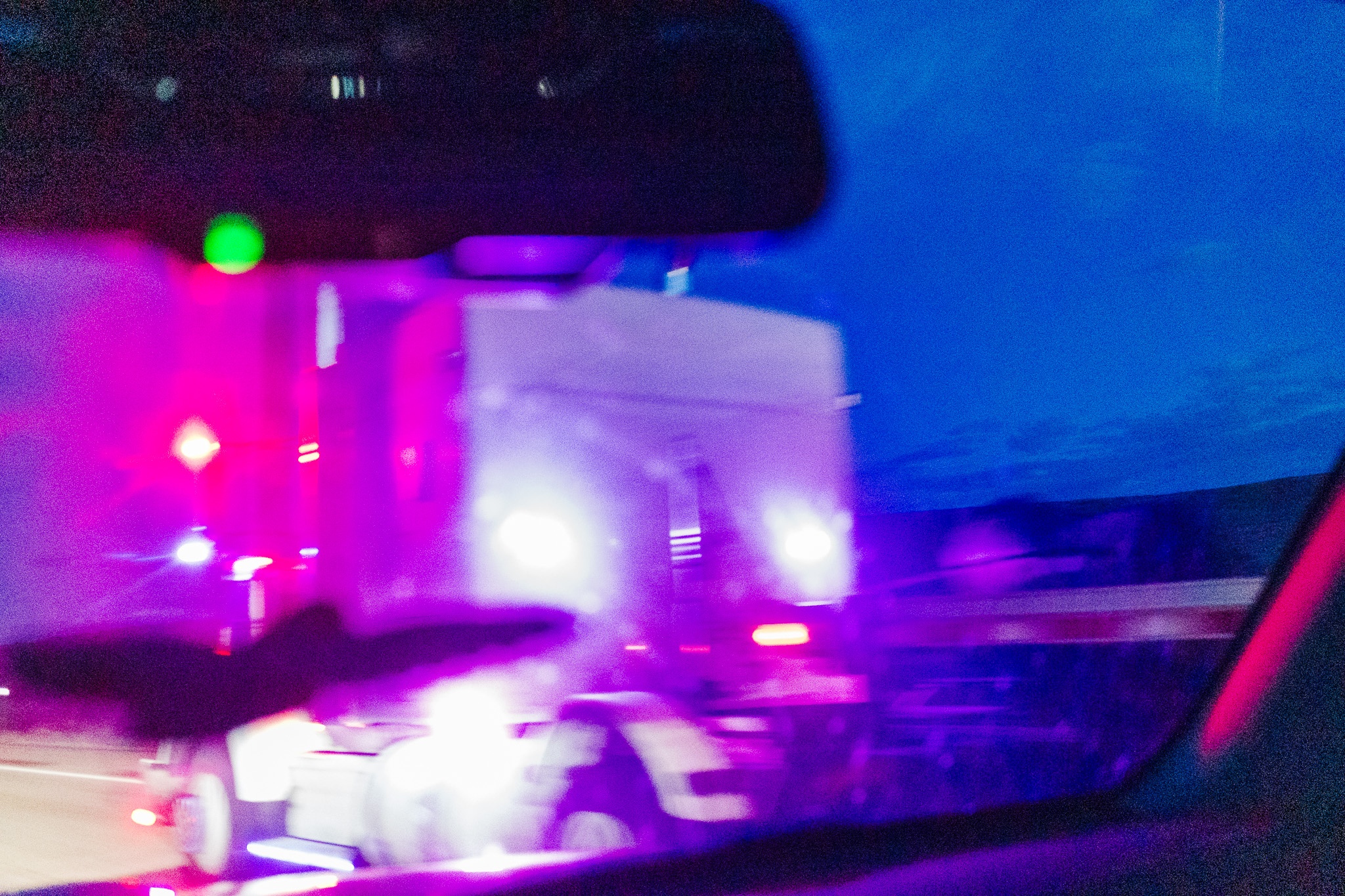 The image captures a blurred scene taken from a vehicle, showcasing a large truck illuminated by vibrant purple and blue lights, likely from emergency vehicles nearby. The setting appears to be nighttime, with a dark sky contributing to the dramatic lighting. The foreground includes part of the viewer's car, suggesting a sense of motion as the truck passes by. The overall composition conveys a sense of urgency or action, amplified by the colorful lights and the blurred effect.