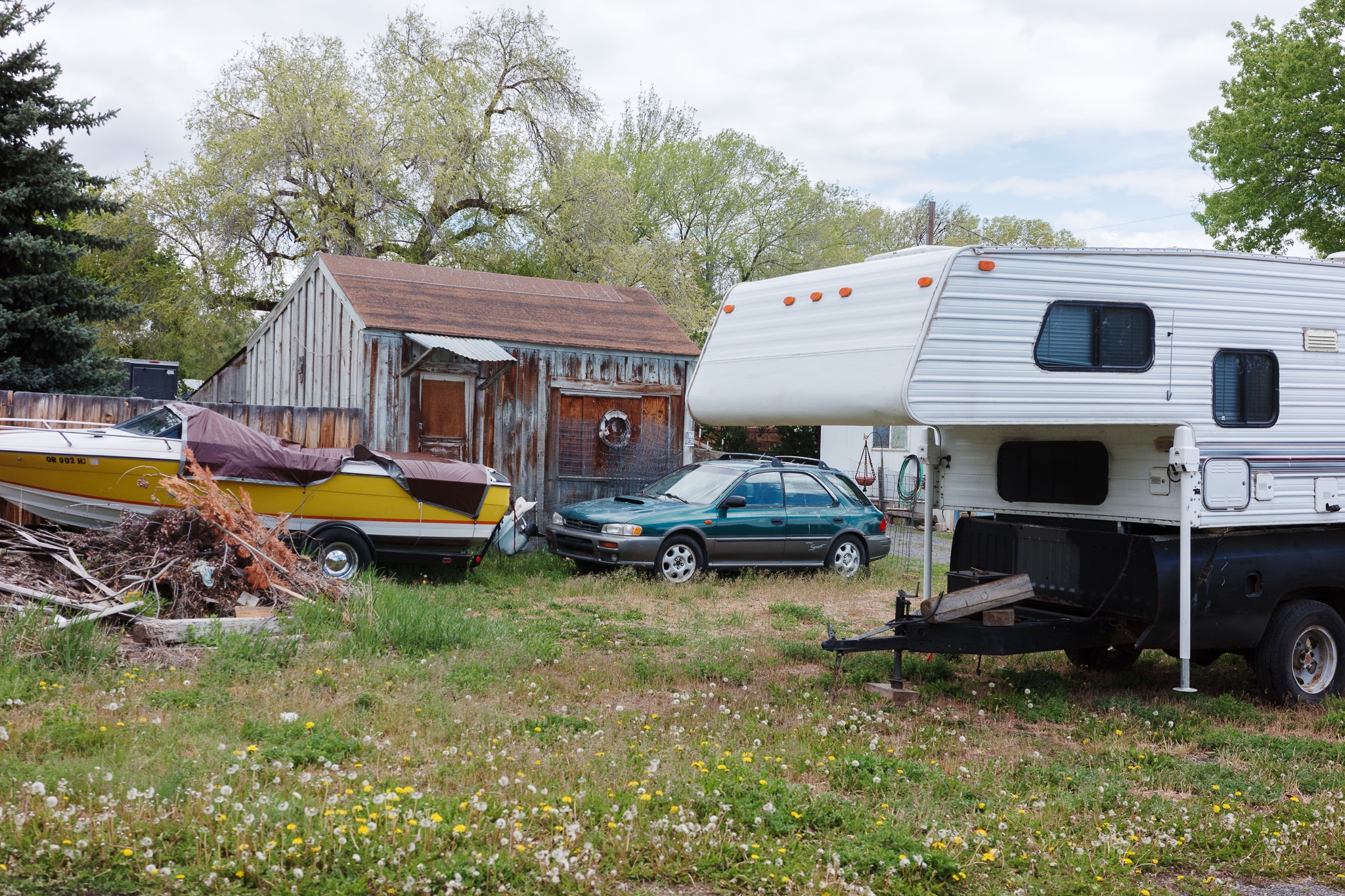 The image depicts a cluttered outdoor setting featuring an assortment of vehicles and structures. In the foreground, a yellow boat partially covered with a tarp sits next to a green car, while a white camper is parked nearby. Behind them is a weathered wooden shed with a rusted roof, surrounded by overgrown grass dotted with wildflowers and dandelions. The scene conveys a sense of rural charm and a relaxed, lived-in atmosphere, with trees providing a backdrop under a partly cloudy sky.