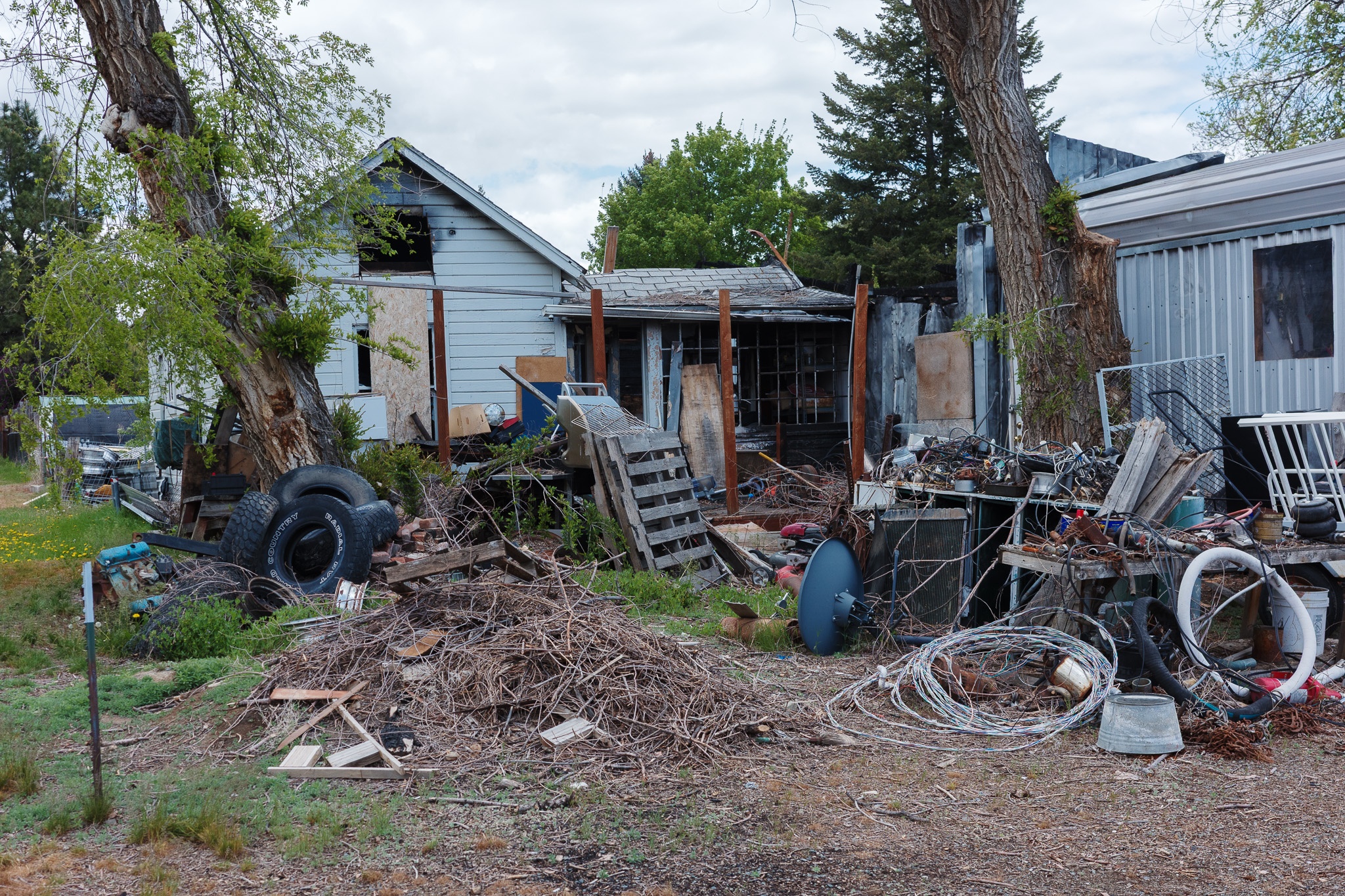 The image depicts a cluttered outdoor scene featuring a dilapidated house with boarded-up windows, surrounded by a chaotic accumulation of debris and discarded items. In the foreground, a pile of tires, wooden pallets, and tangled wires contribute to the disarray, while a large tree stands nearby, its branches reaching out toward the sky. The setting appears neglected, with overgrown grass and scattered debris hinting at a lack of maintenance. The overall atmosphere conveys a sense of abandonment and decay.