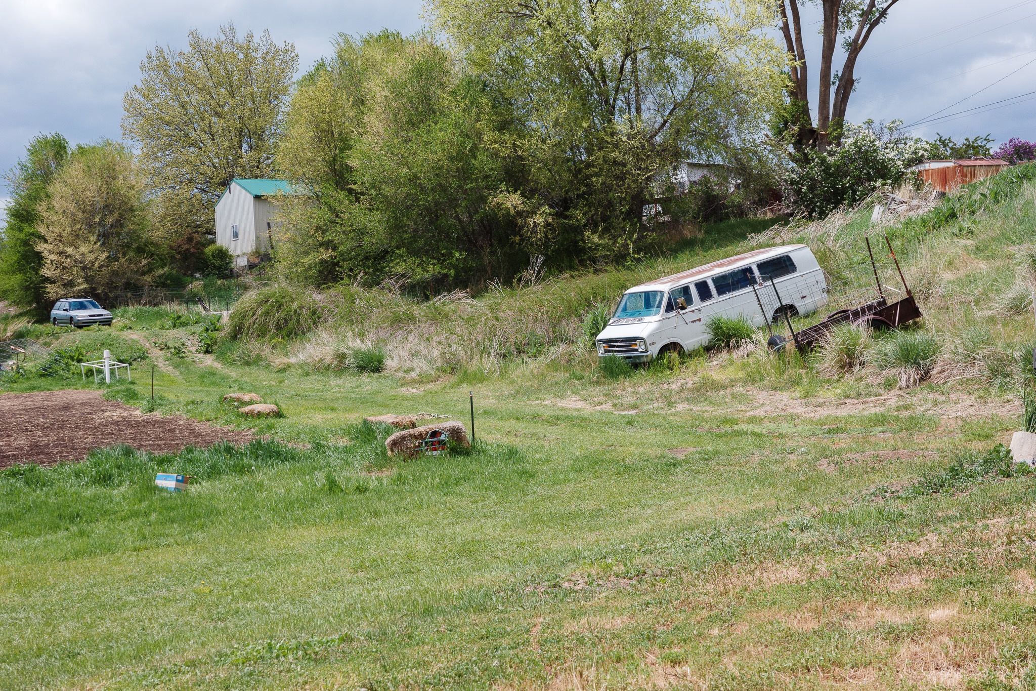 The image depicts a rural scene featuring a grassy area with a dilapidated white van partially hidden among tall grass, suggesting neglect. In the background, a small building with a green roof is nestled among trees that are just beginning to bloom, creating a vibrant spring setting. To the left, a parked car is visible, and scattered throughout the grassy landscape are a few bales of hay and garden plots, adding to the rural ambiance. The cloudy sky above hints at an impending change in weather, contributing