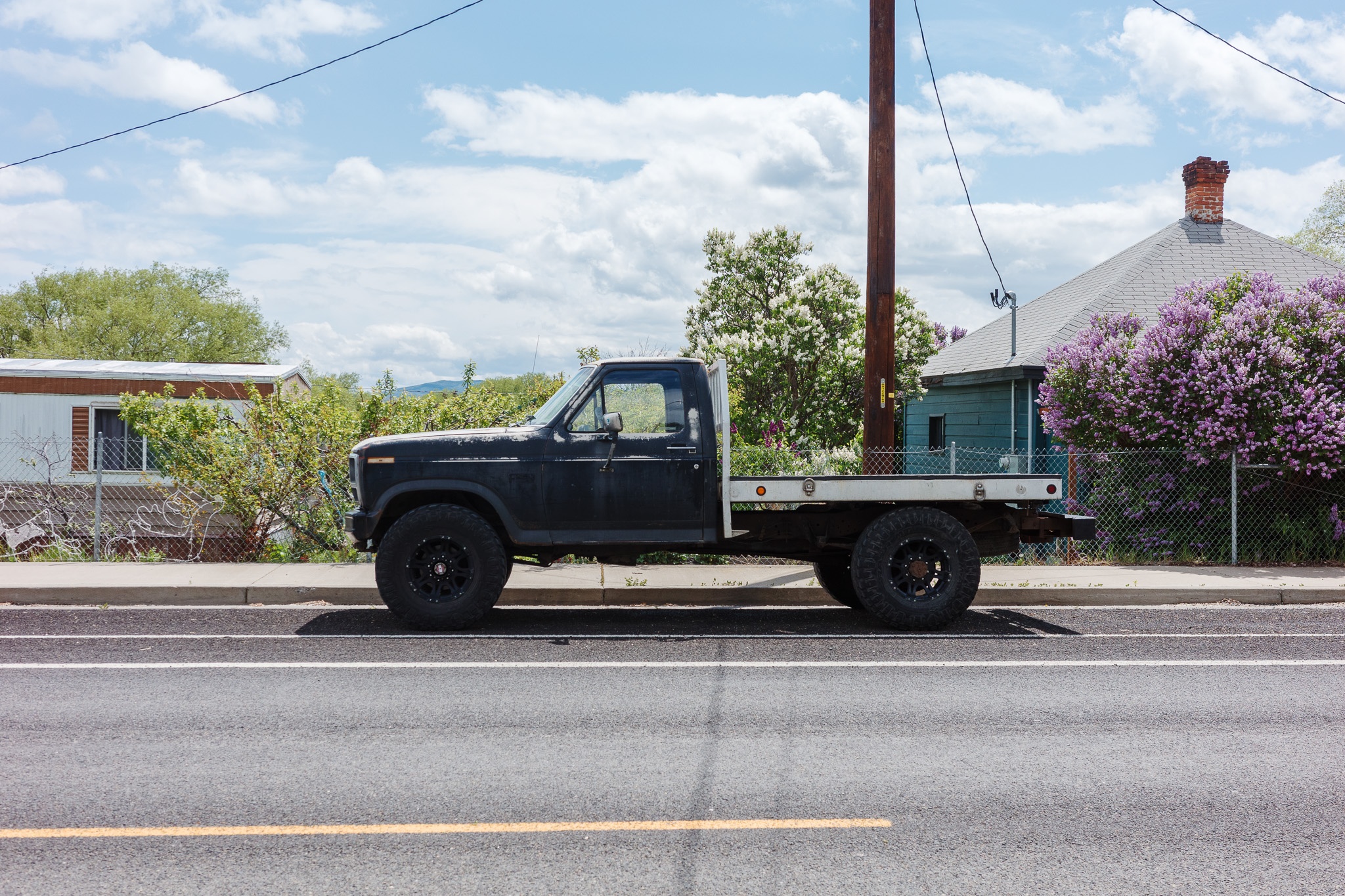 The image features a rugged black pickup truck parked alongside a road, showcasing its weathered appearance with a flatbed design. In the background, there are residential structures, including a house with a gray roof and a blooming purple bush, adding a touch of color to the scene. The sky is partly cloudy, contributing to a bright and open atmosphere, while utility poles and greenery frame the setting, reflecting a rural or suburban environment.