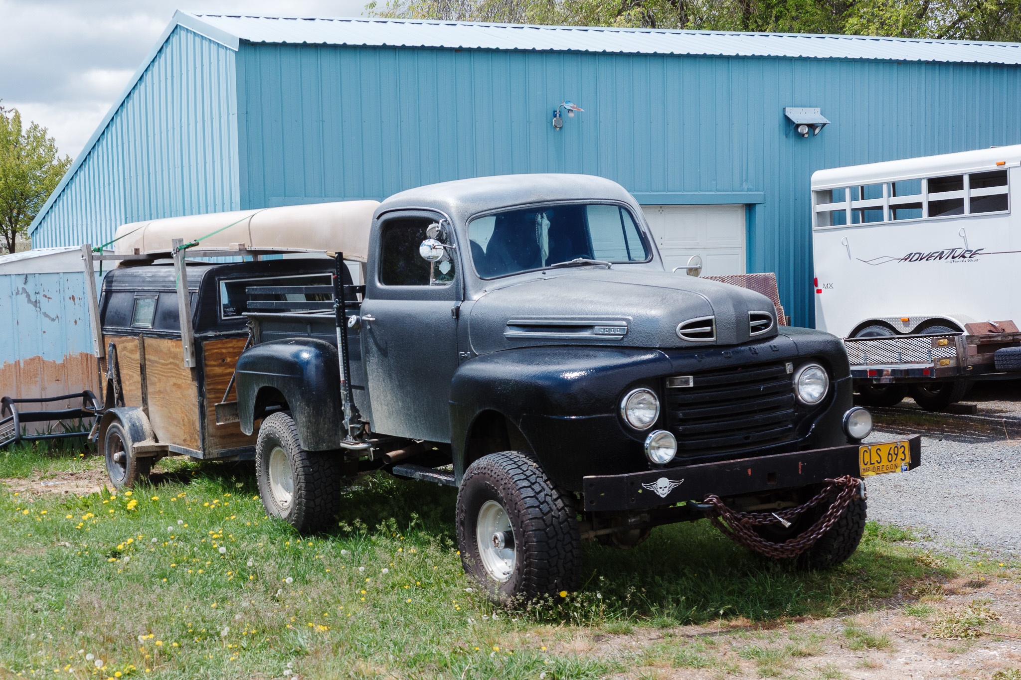 The image depicts a vintage black truck parked in a grassy area, showcasing its rugged appearance with oversized tires and a wooden trailer attached. The truck features a classic design, with distinctive round headlights and a sturdy build. In the background, a blue metal building adds a contrasting visual element, while other structures, including a horse trailer, are partially visible. The scene is set under a partly cloudy sky, contributing to a nostalgic rural atmosphere.