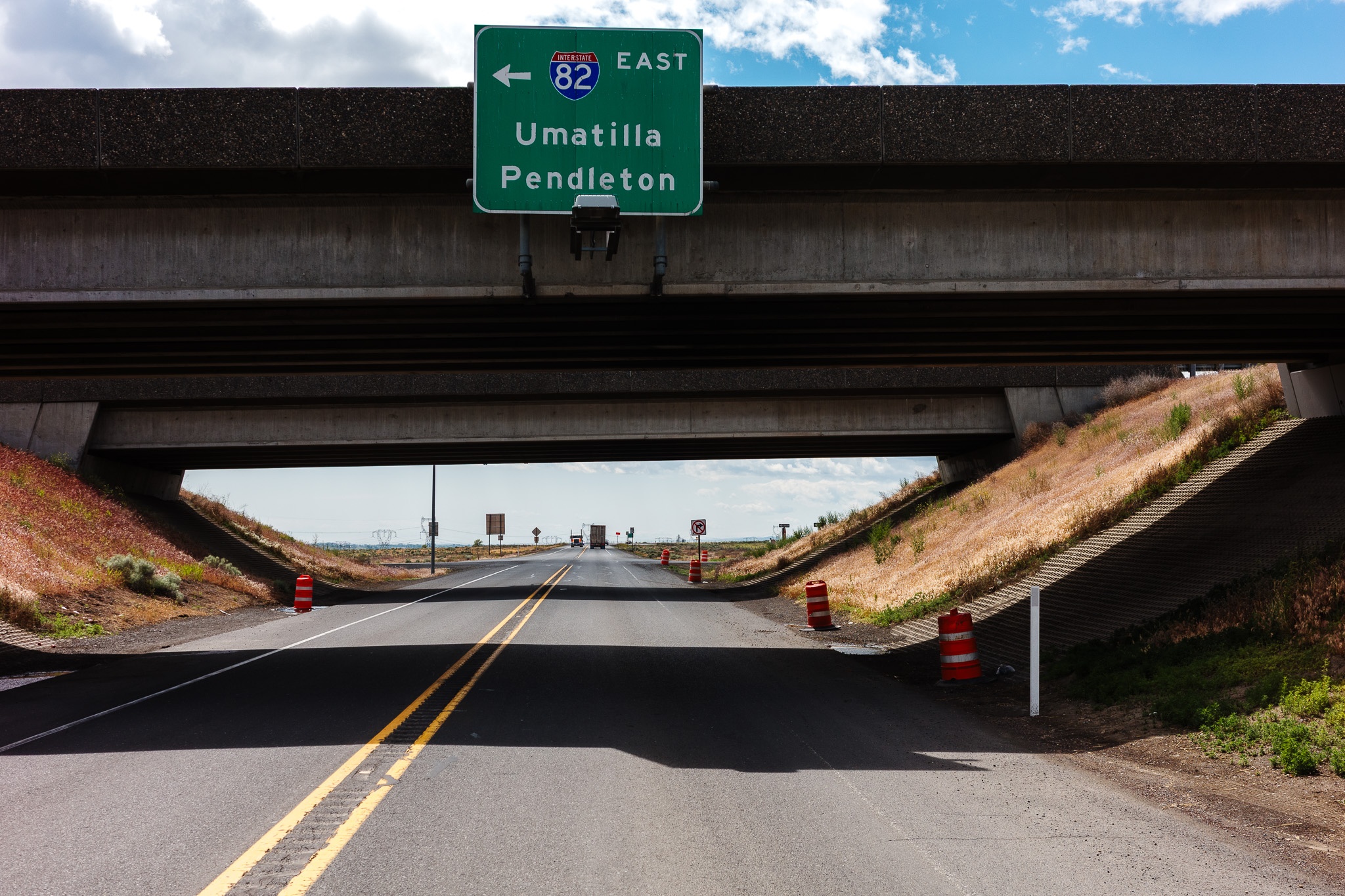 The image depicts a roadway under an overpass, featuring a green highway sign indicating directions to Umatilla and Pendleton. The road appears clear with two traffic cones on the left, while grassy slopes rise on either side. The setting is bright, with a partially cloudy sky overhead, suggesting a sunny day. The overall scene conveys a sense of transportation and movement, framed by the concrete structure of the overpass above.