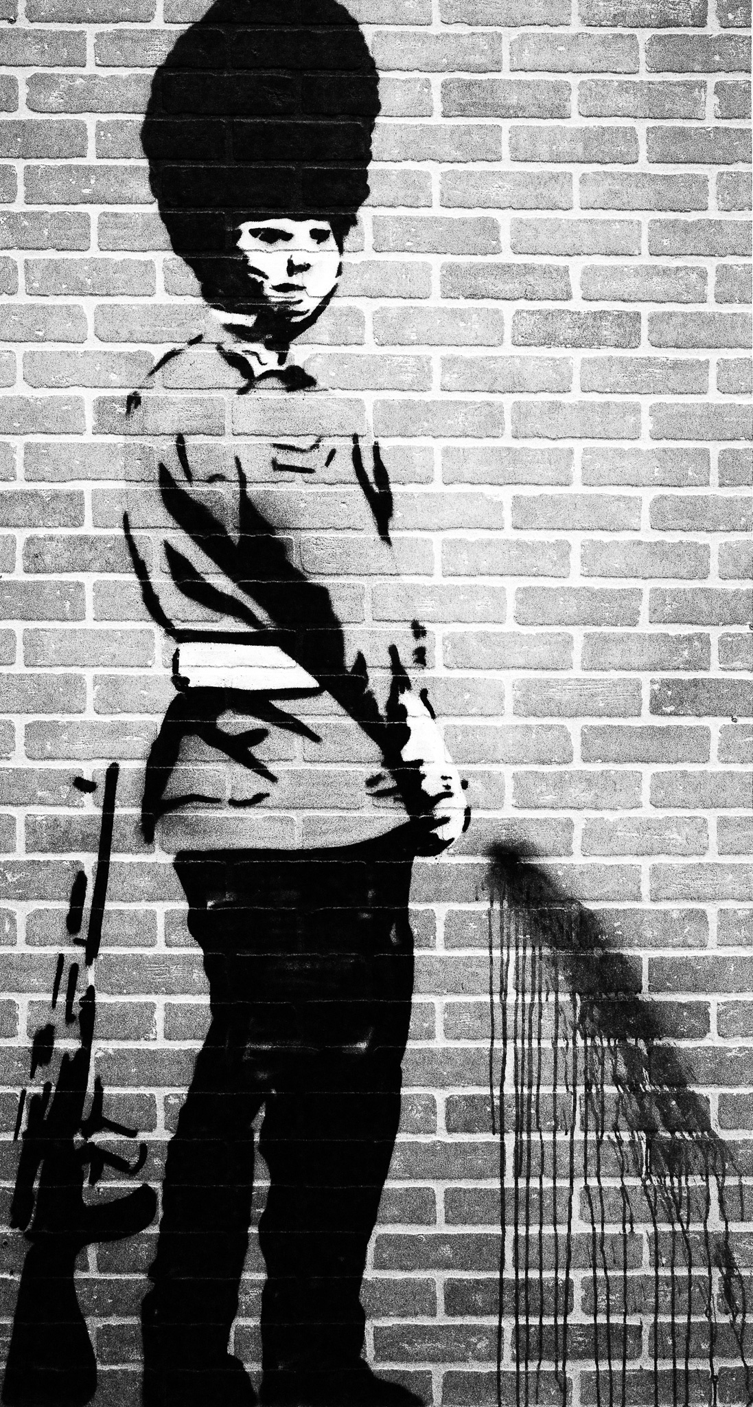 The image features a striking street art depiction of a soldier standing against a brick wall. The soldier, dressed in military attire and wearing a tall fur hat, holds a rifle by his side, exuding a sense of authority. The artwork is rendered in monochrome, emphasizing the contrast between the dark figure and the lighter background of the bricks. Notably, there are drips of paint cascading down from the soldier's hand, adding a dynamic element to the composition and suggesting a sense of rebellion or change
