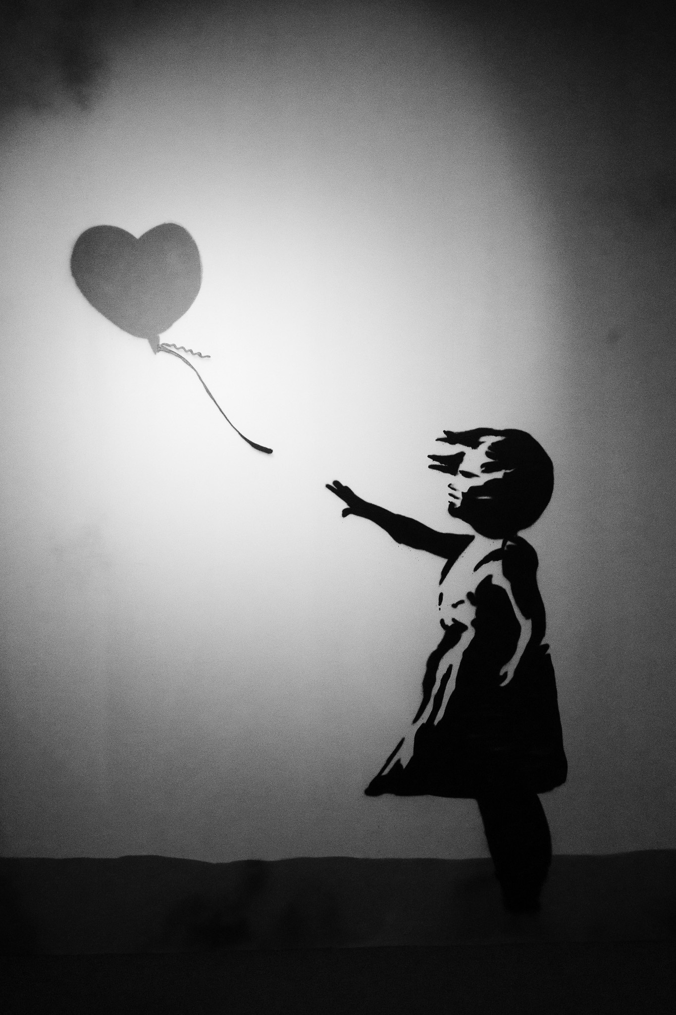 The image features a striking black-and-white mural of a young girl reaching out towards a heart-shaped balloon that floats away from her. The girl, depicted in silhouette, has a windswept hairstyle and is dressed in a simple dress, conveying a sense of innocence and longing. The shadow of the balloon is cast on the wall, enhancing the emotional impact of the scene. The minimalist backdrop adds to the poignancy of the moment, capturing a blend of hope and loss.
