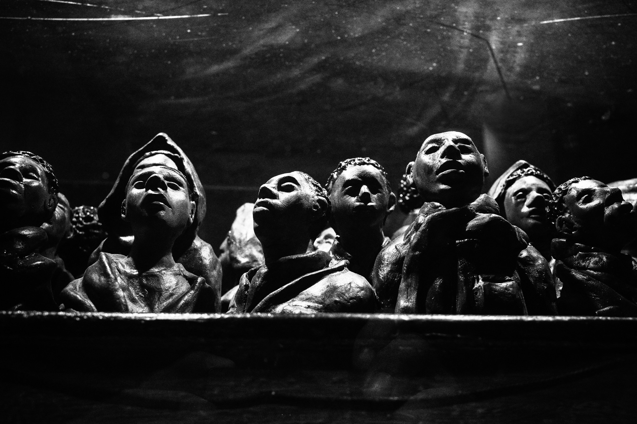 The image features a collection of intricately crafted sculptures, depicting figures with expressive faces, seemingly engaged in a moment of contemplation or reverence. The figures are arranged closely together, their heads tilted slightly upwards, suggesting a shared experience or emotion. The lighting is dramatic, casting shadows that enhance their features and lend a solemn atmosphere to the setting, which appears to be a display case or an exhibition space. The monochromatic color scheme further emphasizes the depth and texture of the sculptures, creating a powerful visual impact