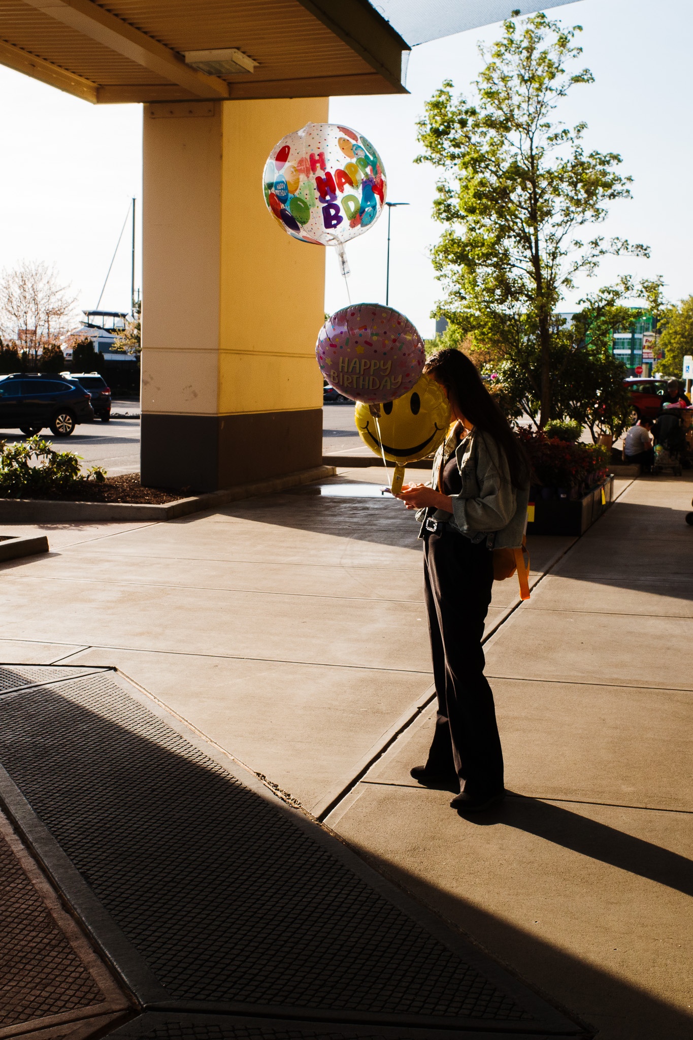 The image captures a young woman standing outside a building, holding three colorful balloons with birthday messages. She appears to be checking her phone while dressed casually in a denim jacket and dark pants. The setting features a sunny day with natural light casting long shadows on the pavement, and various cars parked nearby, hinting at a lively shopping environment. In the background, greenery and a hint of watercraft can be seen, adding to the vibrant atmosphere.