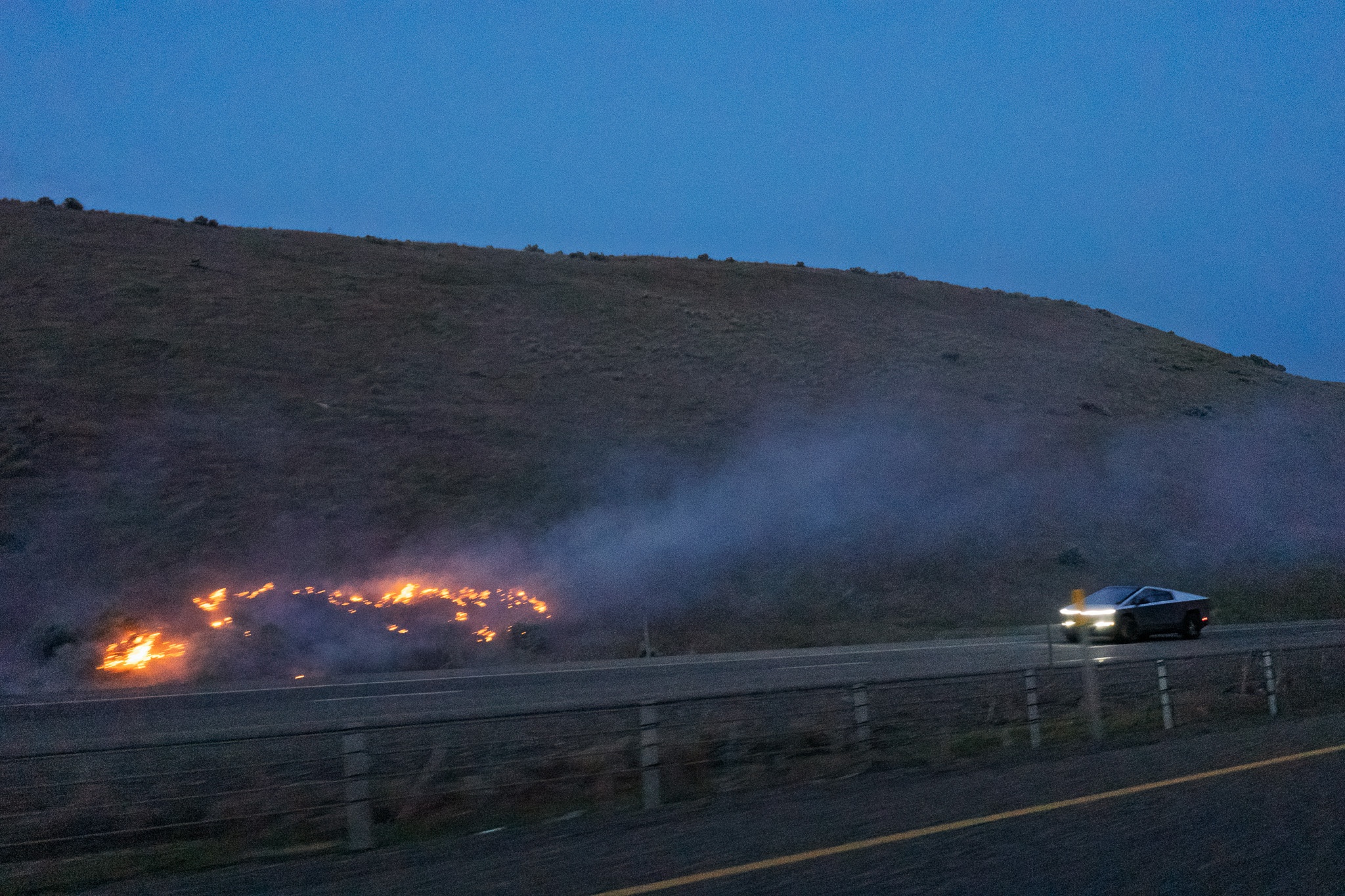 The image depicts a nighttime scene along a roadside where a vehicle is driving past a small brush fire on a hillside. The flames are visible amidst the smoke, creating a stark contrast against the dimly lit landscape. The area is mostly barren with rolling hills in the background, and the car, featuring distinctive headlights, appears to be moving away from the fire. The overall atmosphere conveys a sense of urgency and caution due to the ongoing fire incident.